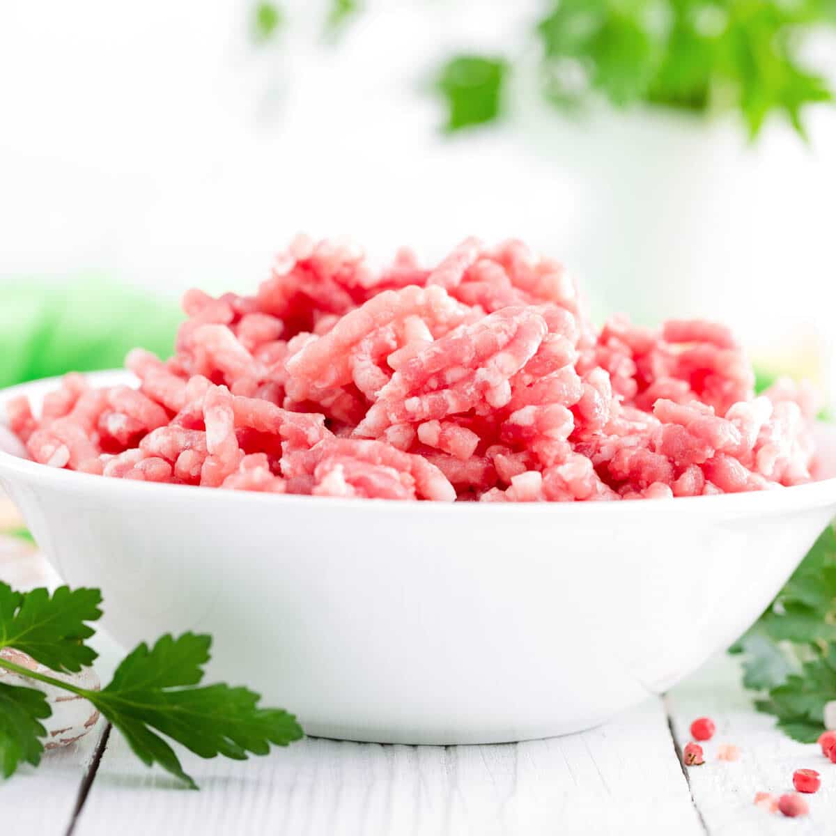 Raw ground pork in a white bowl with sprigs of green herbs.