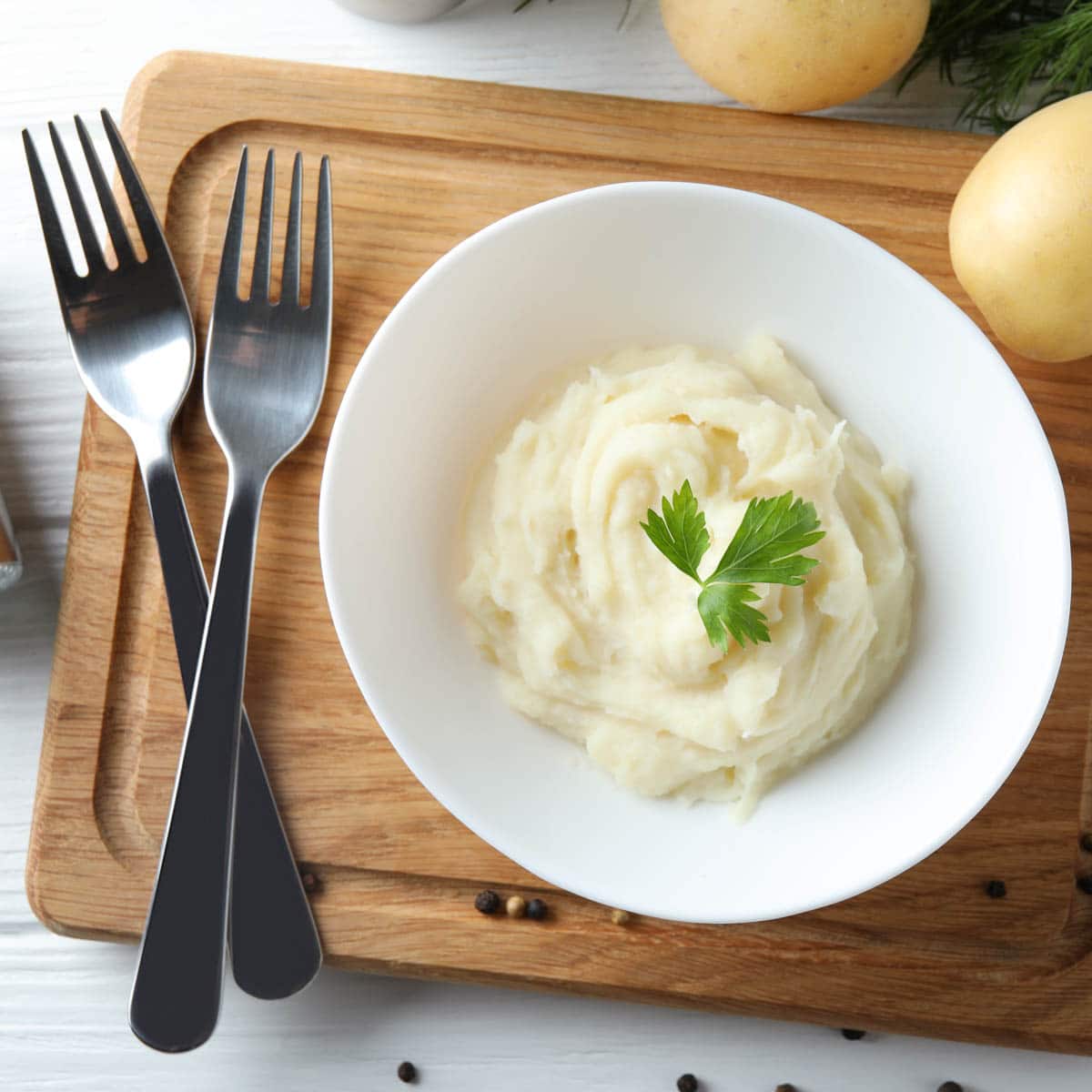 Bowl of mashed potatoes with a sprig of parsley sitting on a cutting board with 2 forks next to it crisscrossed.