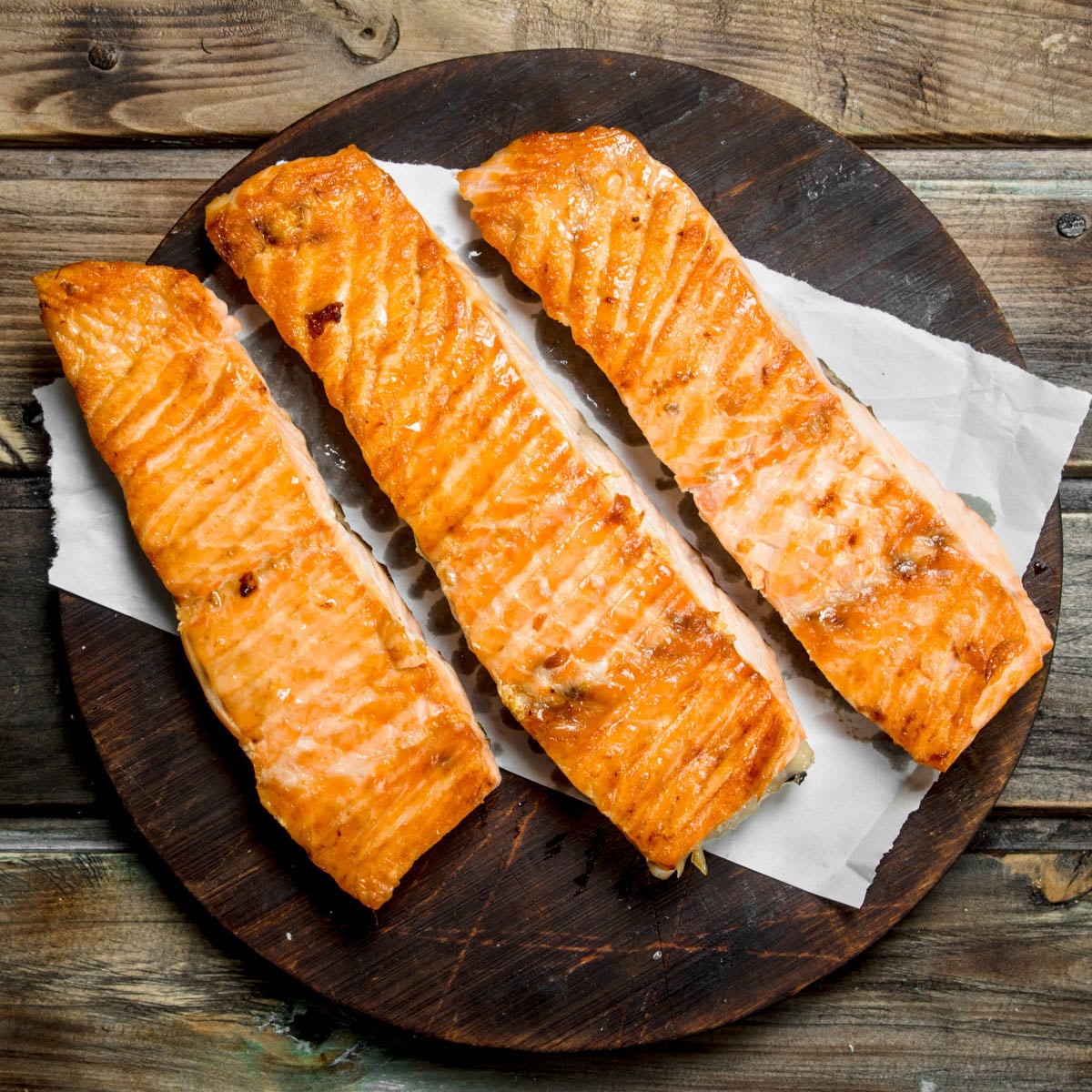 Three cooked salmon filets on a piece of parchment paper on a dark round cutting board with a rustic wooden background