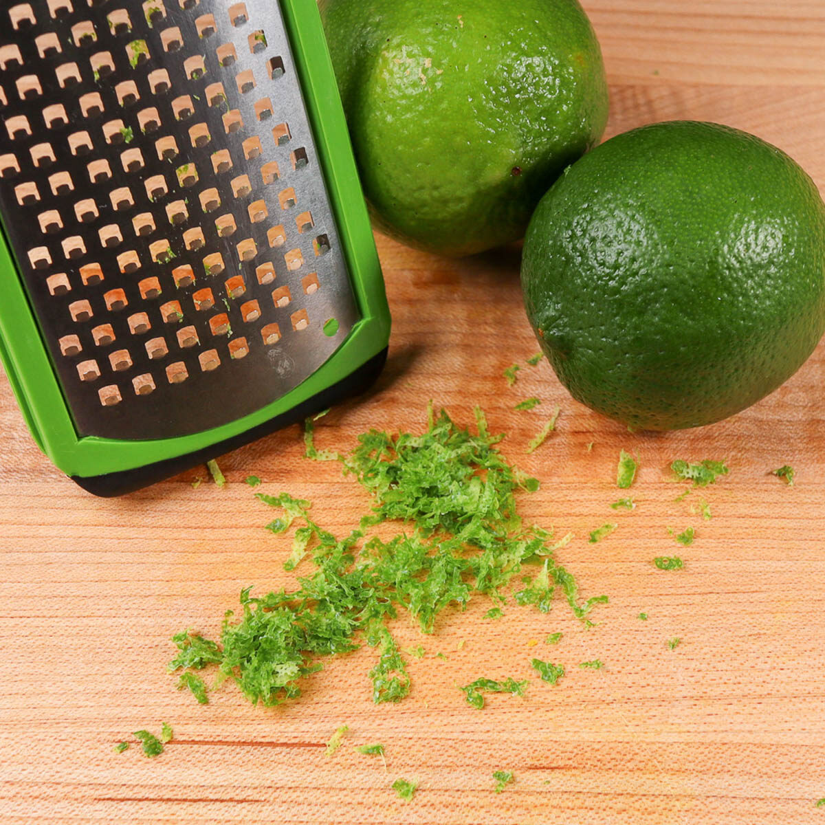Pile of lime zest in front of a mircoplane with two whole limes sitting together off to the side.