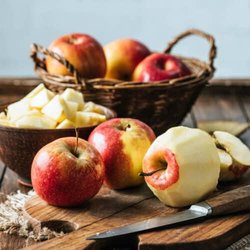Basket of apples with peeled and diced apples in front on a cutting board and a knife.