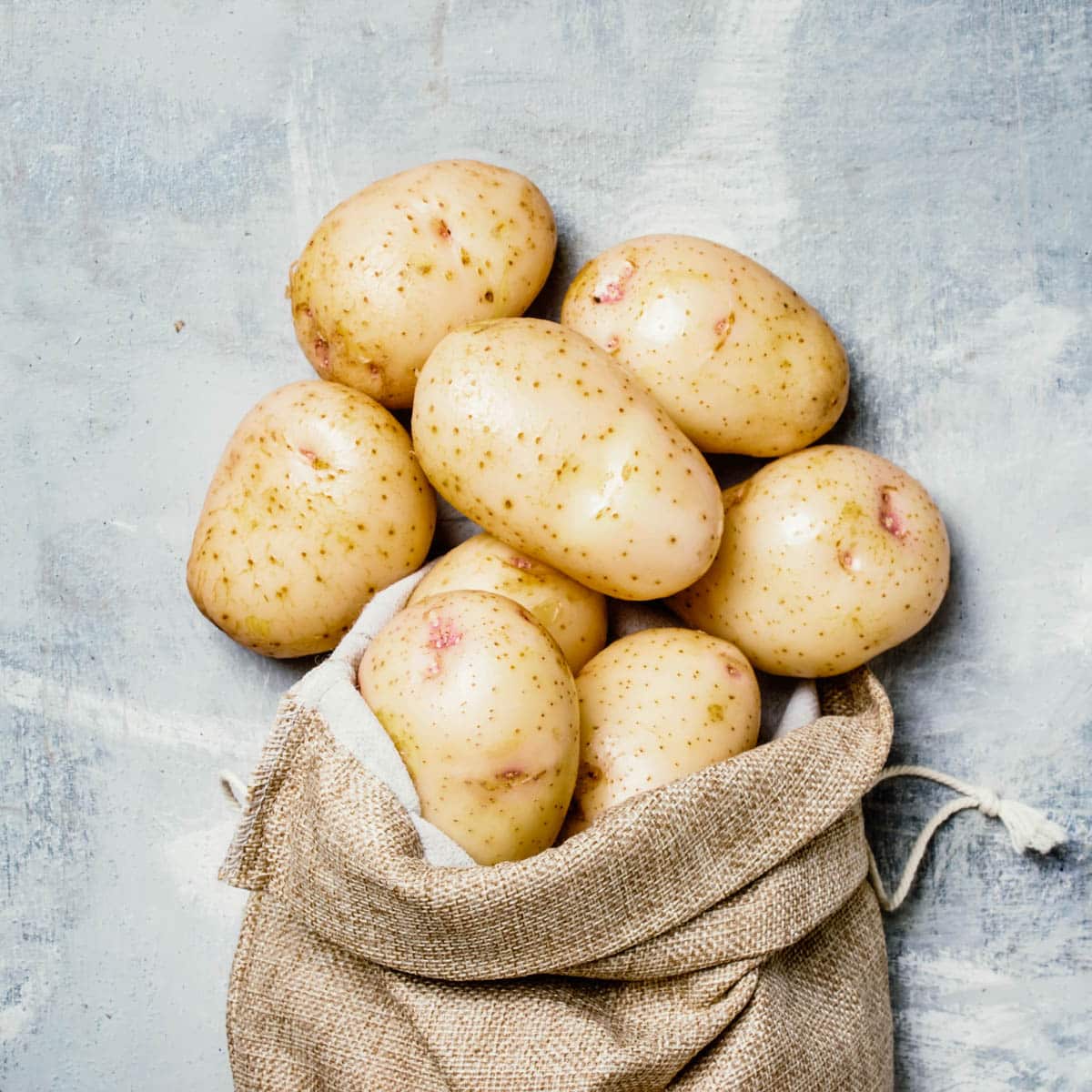 Burlap bag with potatoes spilling out onto a grey background.