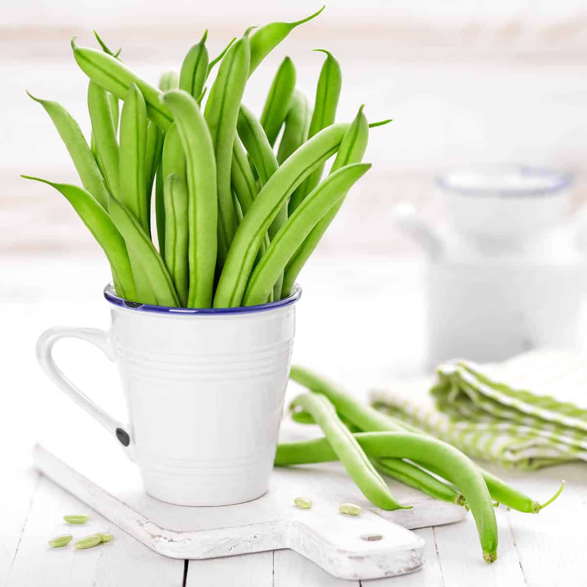 Green beans standing in a white coffee cup on a white cutting board.