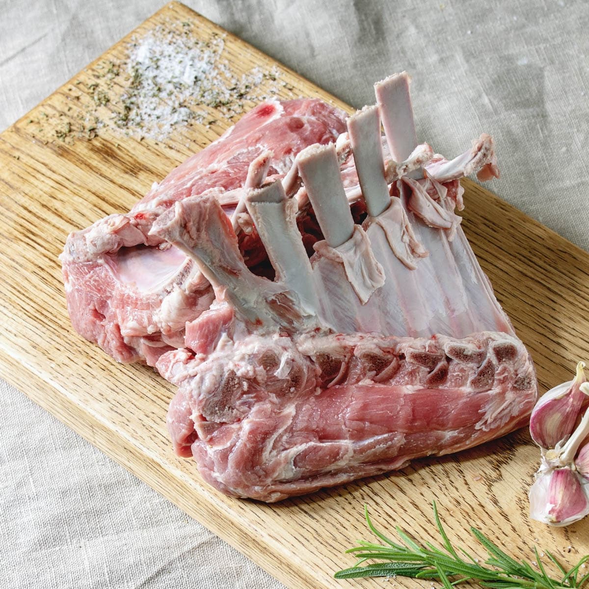 raw rack of lamb on a cutting board with fresh herbs and garlic.