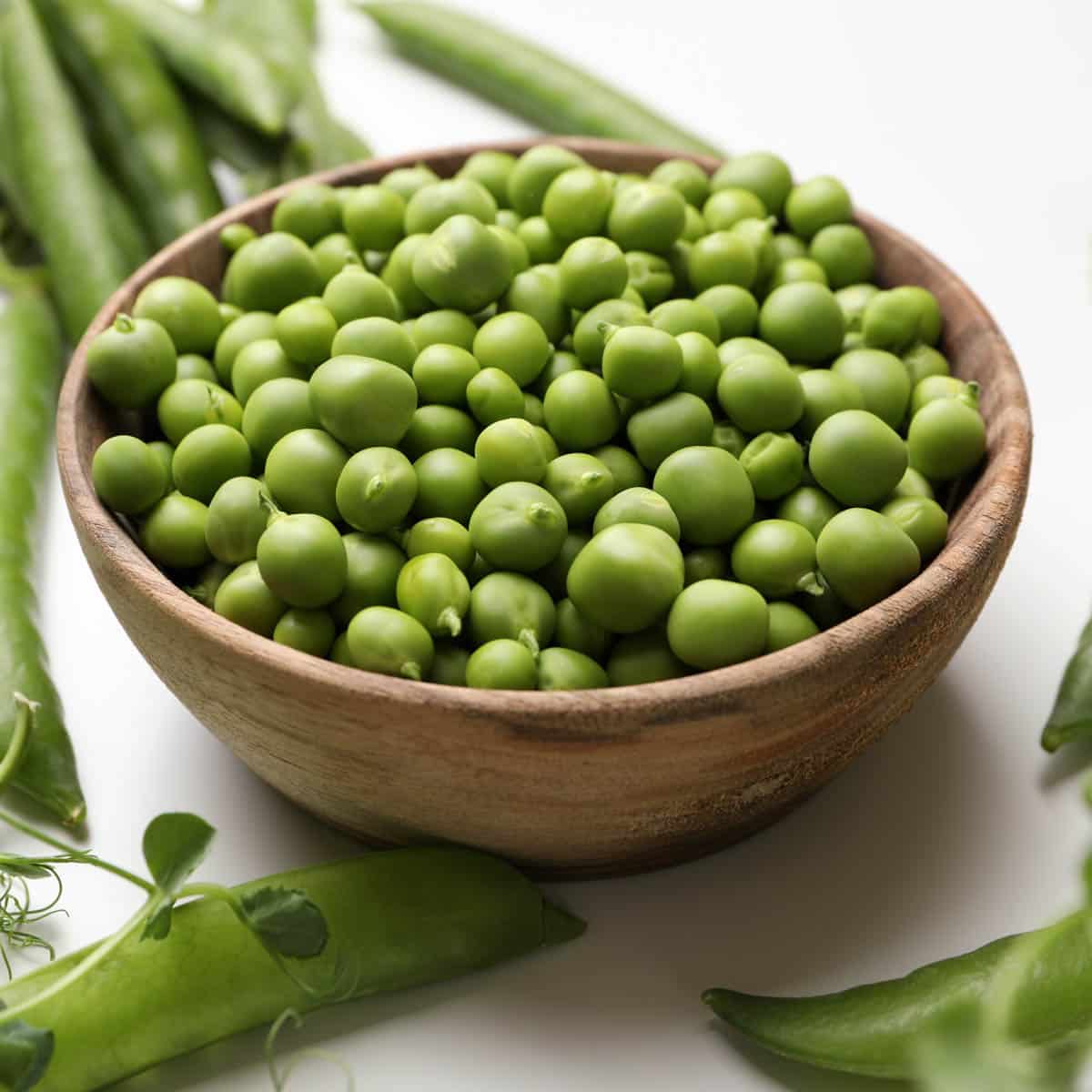 Wooden bowl filled with fresh peas and fresh pods in the background.