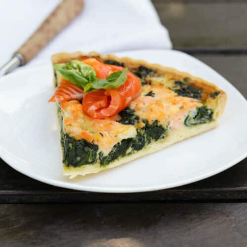A slice of spinach quiche with fresh salmon on top on a white plate.